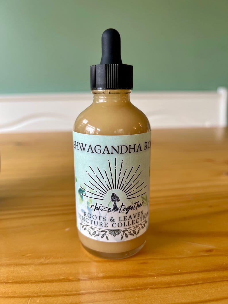 Op de afbeelding: Een heldere glazen fles met een zwarte druppelaar, gevuld met een lichtbruine vloeistof. Het etiket leest "Ashwagandha Roots & Leaves Tincture Collection" met een zonnestraalontwerp en het woord "rhize together". De fles staat op een houten oppervlak.