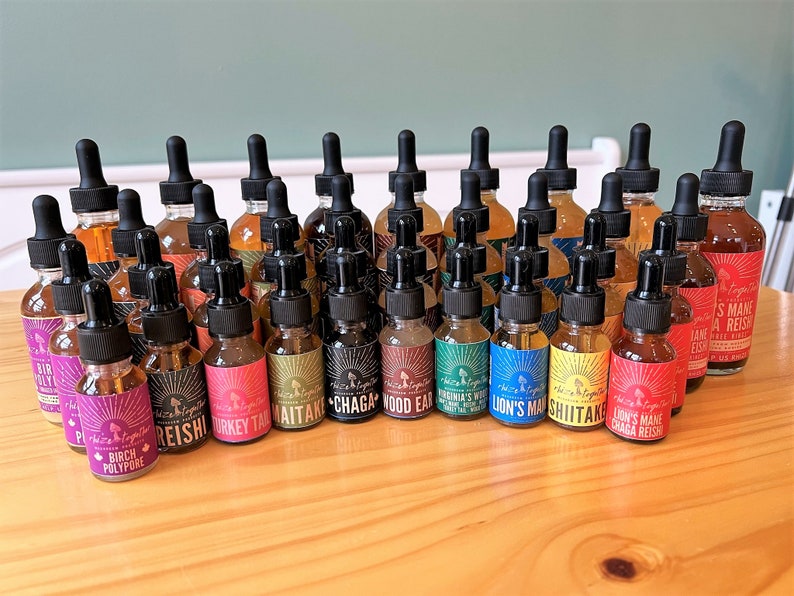 Op de afbeelding: Een verzameling glazen druppelflessen gevuld met amberkleurige vloeistof, elk met een ander label. De labels tonen namen als "Reishi", "Turkey Tail" en "Lion's Mane". De flessen staan op een houten oppervlak.