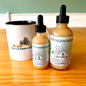 Op de afbeelding: Twee glazen flessen met zwarte druppelaars en een witte mok. De flessen hebben labels met de tekst "Ashwagandha Root" en "Rhize Together Roots & Leaves Tincture Collection". De mok heeft een paddenstoelafbeelding en de tekst "Virginia is for Mushroom Hunters."