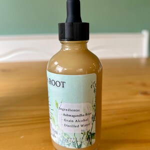 Op de afbeelding: Een heldere glazen fles met een zwarte druppelaar, gevuld met een lichtbruine vloeistof. Het etiket leest "ROOT" en vermeldt ingredi&euml;nten: Ashwagandha Root, Grain Alcohol en Distilled Water. De fles staat op een houten oppervlak.