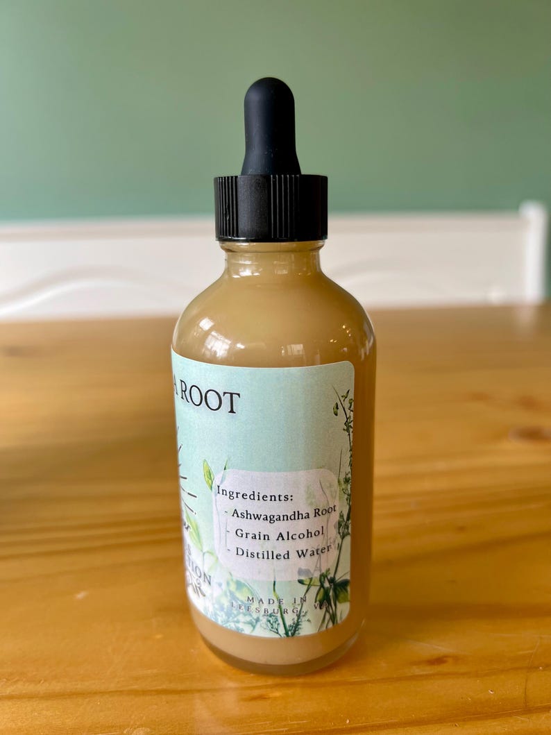 Op de afbeelding: Een heldere glazen fles met een zwarte druppelaar, gevuld met een lichtbruine vloeistof. Het etiket leest "ROOT" en vermeldt ingredi&euml;nten: Ashwagandha Root, Grain Alcohol en Distilled Water. De fles staat op een houten oppervlak.