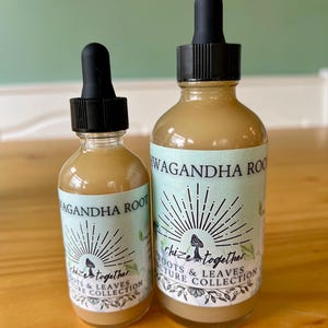 Op de afbeelding: Twee glazen flessen met zwarte druppelaars gevuld met een lichtbruine vloeistof. De etiketten luiden "WAGANDHA ROOT" en "rhize together ROOTS & LEAVES TURE COLLECTION" met een paddenstoelafbeelding en een zonnesterontwerp. De flessen staan op een houten oppervlak.