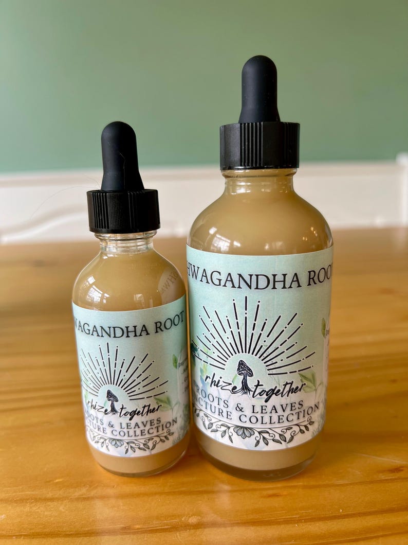 Op de afbeelding: Twee glazen flessen met zwarte druppelaars gevuld met een lichtbruine vloeistof. De etiketten luiden "WAGANDHA ROOT" en "rhize together ROOTS & LEAVES TURE COLLECTION" met een paddenstoelafbeelding en een zonnesterontwerp. De flessen staan op een houten oppervlak.