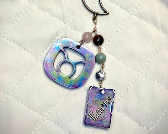 May include: A silver charm with the Taurus zodiac symbol and the word "Taurus" on a silver tag, hanging from a gold crescent moon charm with a chain of beads.