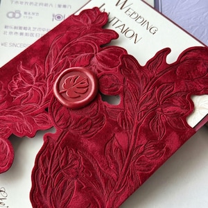 May include: A deep red velvet wedding invitation cover featuring an intricate embossed floral pattern and a circular wax seal with a monstera leaf design. The invitation is partially open, revealing a white card with black printed text.