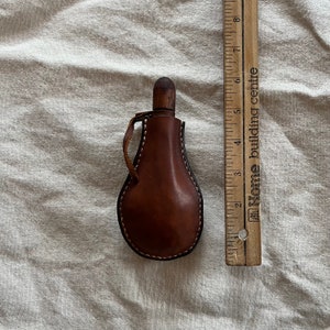 May include: A brown leather flask with a wooden top and a stitched handle. The flask is about 5 inches tall and is sitting on a white fabric surface.