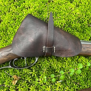 May include: A brown leather gun holster with a strap and buckle. The holster is attached to a wooden gun with a metal trigger.