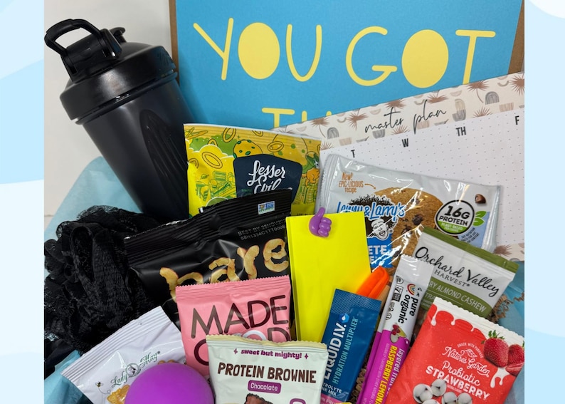 A gift box filled with fitness and wellness items. Includes a black shaker bottle, snacks, protein bars, and a notepad with the text "YOU GOT THIS". The box is an assortment of healthy snacks