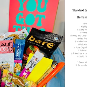 A gift box filled with snacks, a pen, highlighter, sticky note pad, and stress ball. The box includes a variety of packaged snacks, a purple stress ball, and a card with the words "YOU GOT THIS" in teal.