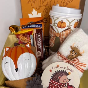 May include: A gift box containing a fox-shaped pumpkin, a travel mug with a pumpkin pattern, a pair of white fuzzy socks with a highland cow patch, a Ghirardelli chocolate square, a MadeGood granola bar, a gold spoon, and a round sign reading Love ya a latte.
