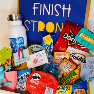 May include: A gift basket filled with snacks, drinks, and small gifts. Includes a white water bottle, chips, fruit snacks, lip balm, and a blue figurine. The words "FINISH STRONG" are displayed on a blue background.
