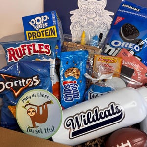 May include: A gift box containing various snacks including Pop-Tarts, Ruffles, Doritos, Chips Ahoy, and Oreo Minis. A white water bottle with the text Wildcats, a round sticker with a sloth and the text Hang in there... You got this!, and a small football are included.
