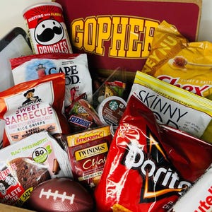May include: A gift box filled with assorted snacks including a Pringles can, Chex Mix, SkinnyPop, Doritos, Nutella, Quaker rice crisps, Lenny & Larry's cookie, Twinings tea, Emergen-C, a Snickers bar, and a small brown football, with a GOPHER sign in the back.