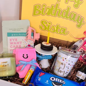May include: A birthday gift box containing a yellow card with the text Happy Birthday Sista, a green candle labeled it's your birthday, pink socks with a smiley face, a sheet mask, a hair clip, a mini cake with a candle, a glass cup, and Oreo cookies.