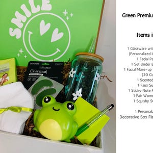May include: A green gift box filled with self-care items. Includes a glass with a bamboo lid, facial mask, eye patches, makeup remover wipes, a scented sachet, a faux succulent, a sticky note pad with a pen, socks, and a squishy frog.