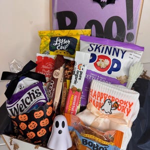 以下が含まれることがあります： SkinnyPopのポップコーン、Welch'sのフルーツスナック、Bobo'sのパンプキンスパイスバー、Pureのフルーツバーなどのスナックが入ったギフトボックス。お化けの柄の白い靴下、小さなお化けのフィギュア、黒いコウモリの切り抜きが含まれています。