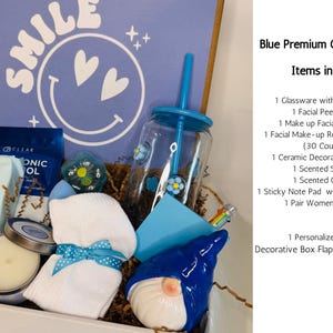 May include: A blue gift box with a variety of items, including a ceramic gnome, a candle, a tumbler with a straw, and a pair of socks. The box has a blue lid with the word "SMILE" and a smiley face design.