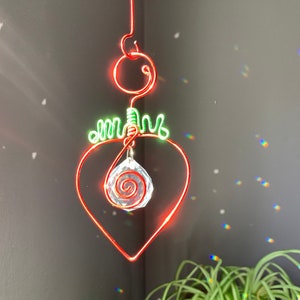 May include: A red wire-wrapped heart with a green wire-wrapped stem and leaves. A clear crystal with a spiral design hangs from the center of the heart.