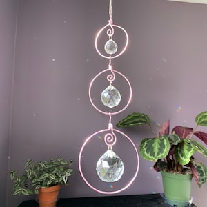 May include: A pink metal wire crystal suncatcher with three clear crystal prisms hanging from it. The suncatcher is hanging from the ceiling and is surrounded by plants.