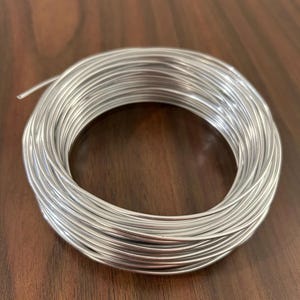 May include: A coil of shiny, silver-colored wire. The wire is tightly wound, forming a circular shape. The wire appears to be made of metal and is likely used for crafting or jewelry making. The background is a dark brown wooden surface.
