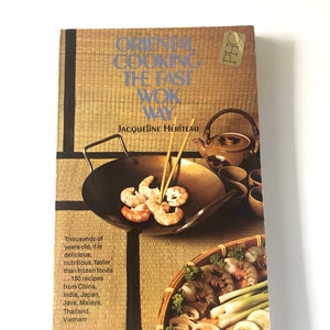 May include: A cookbook titled "Oriental Cooking The Fast Wok Way" by Jacqueline Hériteau. The cover shows a wok with prawns and chopsticks, a teapot, and a basket of vegetables. The book's theme is Asian cuisine.