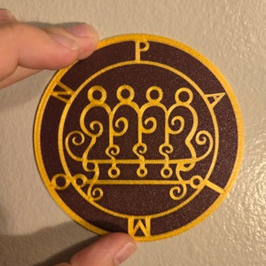 May include: A round, brown patch with a gold embroidered sigil. The sigil is a complex design with a series of circles, swirls, and lines. The patch has the letters "N", "P", "O", "W", and "A" around the edge.