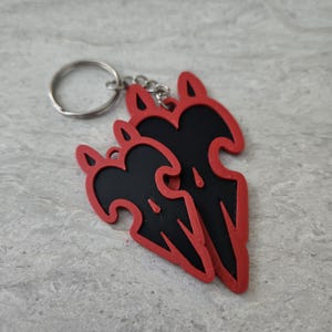 May include: A black and red keychain with a stylized design of three wings. The keychain is attached to a silver metal ring.