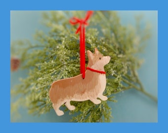 May include: A flat, tan and white Corgi dog ornament with a painted finish. The dog figure has a red ribbon tied around its neck and a matching red ribbon loop at the top for hanging. The ornament is shaped like a side profile of a Corgi.