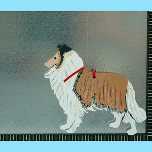 4” by 4” wooden cutout of a Collie in silhouette with a magnet on one side. The Collie is realistically hand painted in shades of golden brown, white and black and wears a thin red satin collar. There is a small hole drilled for a tag or ribbon.