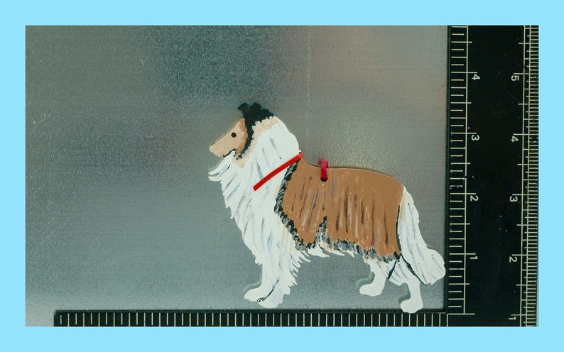 4” by 4” wooden cutout of a Collie in silhouette with a magnet on one side. The Collie is realistically hand painted in shades of golden brown, white and black and wears a thin red satin collar. There is a small hole drilled for a tag or ribbon.