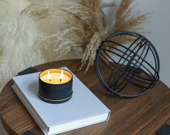 May include: A black candle in a tin container with a gold rim, burning with two flames, sits on a gray book on a wooden table. A black metal orb with a geometric design is in the background.