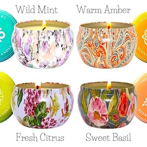 May include: Four scented candles in decorative tins. The candles are labeled "Wild Mint", "Warm Amber", "Fresh Citrus", and "Sweet Basil". Each tin has a unique floral or patterned design. The candles are lit, with flames visible.