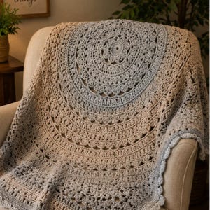 May include: A circular crochet blanket featuring a mandala pattern with a gradient color transition from light blue at the center to beige and grey tones toward the outer edges, showcasing intricate stitch work and a scalloped decorative border.