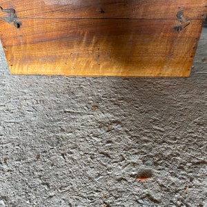 May include: A wooden box with a hinged lid and metal clasps. The box is made of light brown wood with visible grain and some darker areas. The box is sitting on a rough, grey surface.