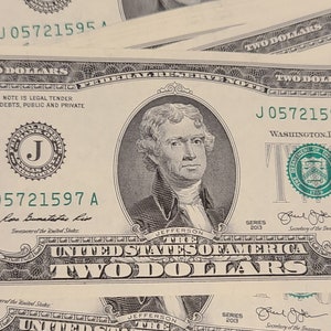 Pode incluir: Uma nota de dois dólares com o número de série J05721597 A. A nota apresenta um retrato de Thomas Jefferson e as palavras "United States of America" e "Two Dollars".