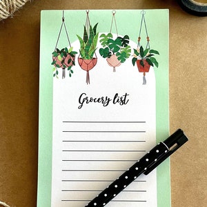 May include: A green grocery list notepad with a hanging plant design. The notepad has a white background with black lines and the words "Grocery List" printed in black. A black pen with white polka dots is lying on the notepad.