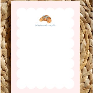 May include: A pink notepad with a scalloped edge and a white interior. The notepad features a small illustration of a croissant with a blue ribbon and the text "Un Croissant, s'il vous plaît".