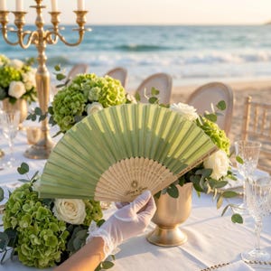 May include: A light green folding fan with a wooden handle is held in front of a table setting. The fan has the text "Cheers to another great year!" and "Honey & Love" printed on it. The table is set with flowers and candles.