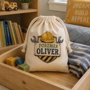 May include: A cream-colored drawstring bag featuring a graphic of a yellow construction helmet, two crossed wrenches, and a shield with black and yellow hazard stripes. The text reads FOREMAN OLIVER in bold, dark blue and green lettering.