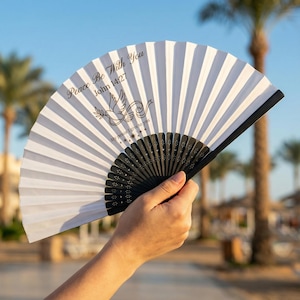 May include: A white and black folding fan with the text "Peace Be With You John 14:27" and a dove graphic. The fan is held open, displaying its pleated design. The fan's handle is black.