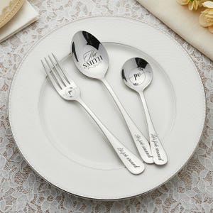 May include: A stainless steel fork, spoon, and teaspoon set on a white plate. The spoon is engraved with "The SMITH", and the fork and spoons have "po" and "Let's fork around" text. The plate has a silver rim.