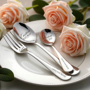 May include: A silver-colored fork, spoon, and teaspoon set on a white plate, with the words "Custom text here" engraved on each piece. Peach-colored roses are in the background, creating a romantic setting.