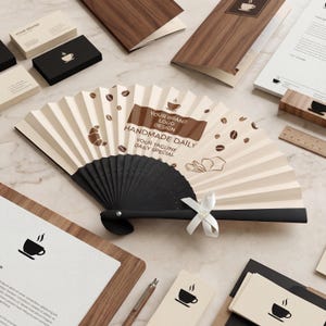 May include: A folded paper fan with a black handle and a white ribbon. The fan has a coffee theme with coffee bean and croissant illustrations. The text on the fan reads "Your Brand Logo Design Handmade Daily Your Tagline Daily Special."