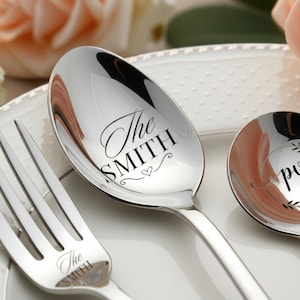 Può includere: Primo piano di un cucchiaio, una forchetta e un piattino d'argento. Il cucchiaio e la forchetta sono incisi con "The SMITH" in elegante scrittura. Un piccolo cuore è disegnato sotto il nome sul cucchiaio. Il piatto ha un'iscrizione parziale. Le posate sono su un piatto bianco.