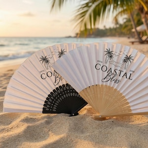 May include: Two open paper fans on a sandy beach. One fan is black, the other is natural wood. Both have white paper with a palm tree design and the words "Coastal Bliss" printed on them. The background shows the ocean and palm trees.