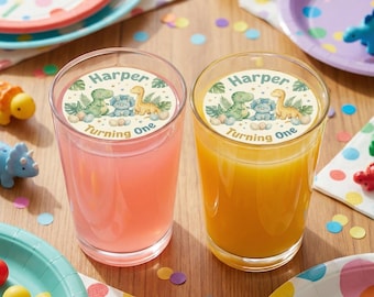 May include: Two clear glasses filled with pink and orange liquid, each with a circular sticker that reads "Harper Turning One" and features dinosaur illustrations. Party plates and confetti are scattered around.