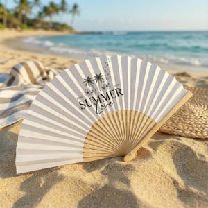 May include: A white paper fan with a wooden handle, resting on sand. The fan features vertical stripes and the words "SUMMER Love" with palm tree and floral graphics. The background shows a beach scene with ocean and sky.
