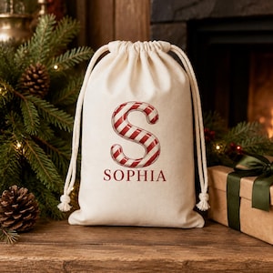 May include: A cream-colored fabric drawstring bag featuring a large letter S designed with red and white candy cane stripes. Below the letter, the name SOPHIA is printed in a dark red serif font. The bag has thick white rope drawstrings.