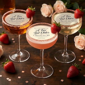 Könnte beinhalten: Drei Cocktailgläser mit Zuckerrand, jedes mit einem Getränk in einer anderen Farbe gefüllt. Der Text "A Tini Bit Older Cheers to 50" ist auf jedem Glas aufgedruckt. Frische Erdbeeren garnieren jedes Glas. Rosen und Konfetti im Hintergrund.