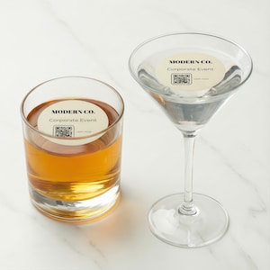 Könnte beinhalten: Zwei klare Gläser, eines ein Whiskeyglas mit bernsteinfarbener Flüssigkeit und das andere ein Martiniglas mit klarer Flüssigkeit. Beide Gläser haben einen runden Aufkleber mit der Aufschrift "MODERN CO. Corporate Event" und einem QR-Code.